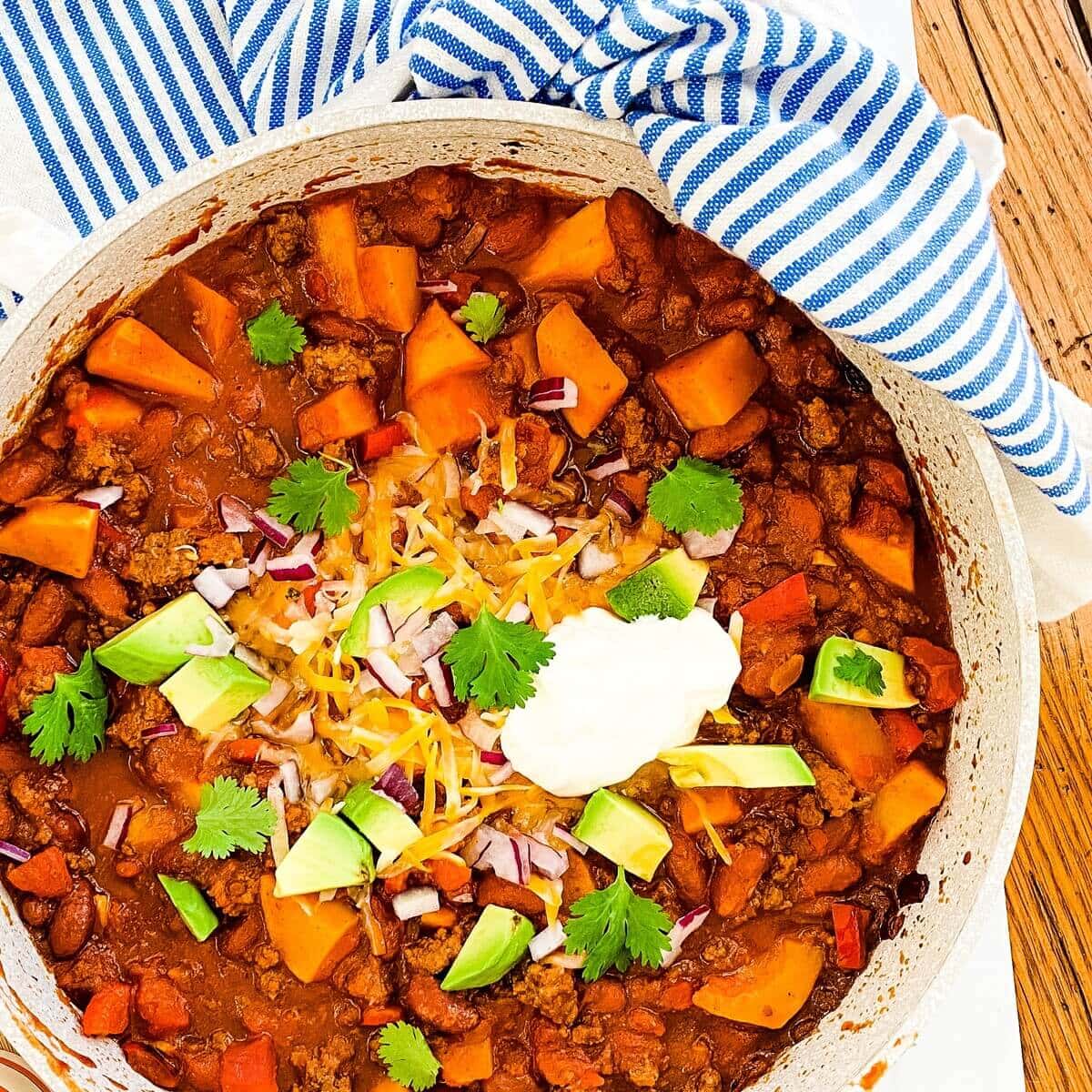 Ground beef and butternut squash chili in a large pot topped with cheese, avocado sour cream and cilantro.