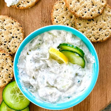 A bowl of tzatziki sauce with crackers and cucumber slices/