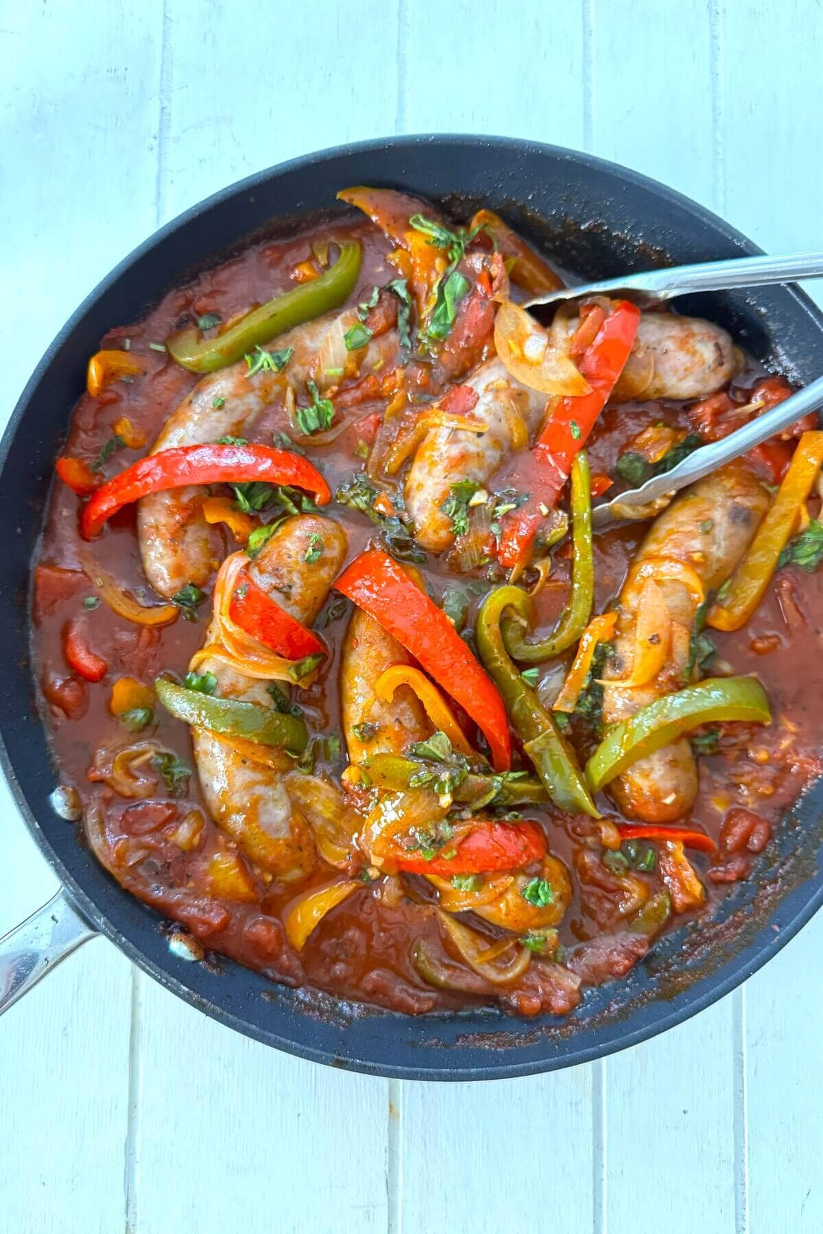A skillet of Italian sausage and peppers with tongs.